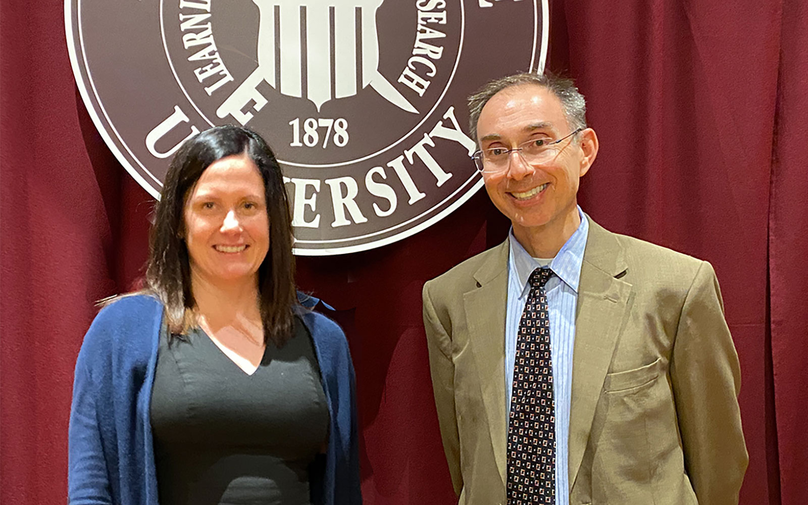 Dr. Ray Uzwyshyn stands with Dr. Lis Pankl in front of a maroon drape background with the MSU seal.