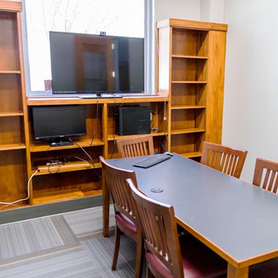 A small meeting room with a video monitor on a bookshelf and a table with chairs.