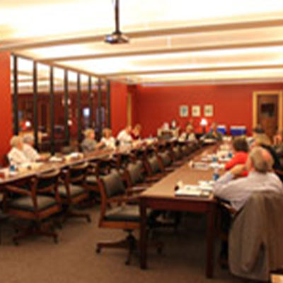 Meeting attendees sit along the two conference tables of a large meeting room.