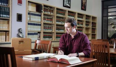 A graduate student takes notes at a table in a research library.