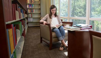 Student sitting and reading next to large picture windows and book shelves.